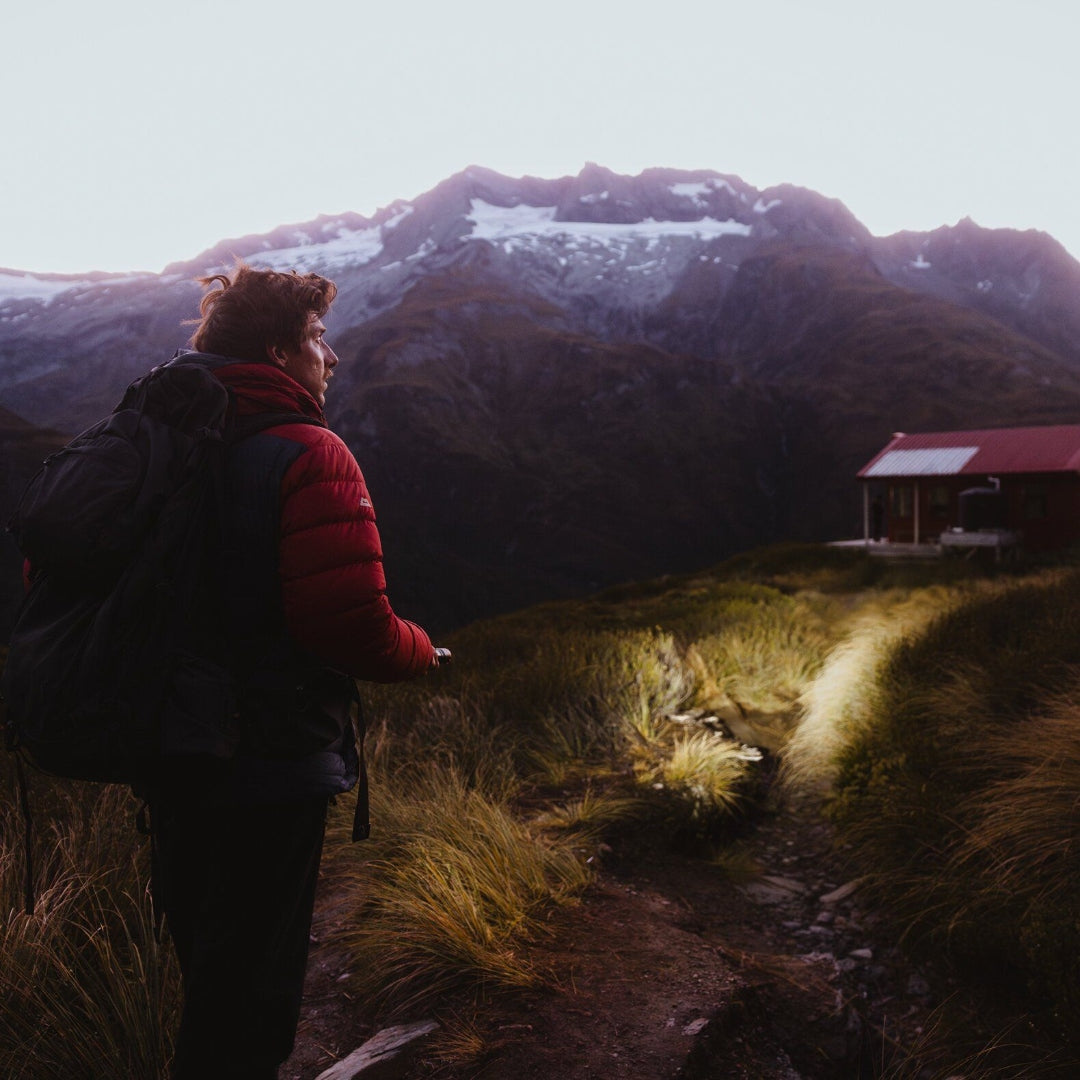Hiker with red jacket and large backpack on a mountain trail at dusk; cabin in distance Ledlenser P7R Signature Torch