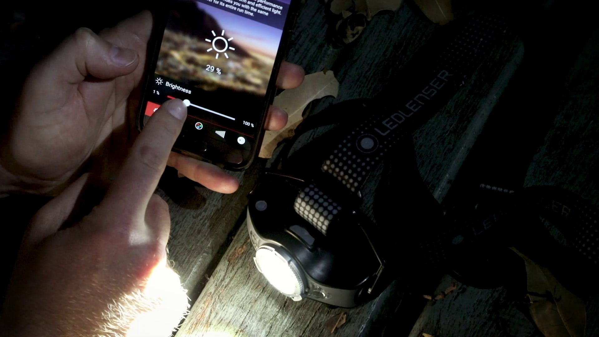 Ledlenser MH11 Blue Outdoor Headlamp on a wooden surface with a person adjusting brightness on a nearby smartphone screen