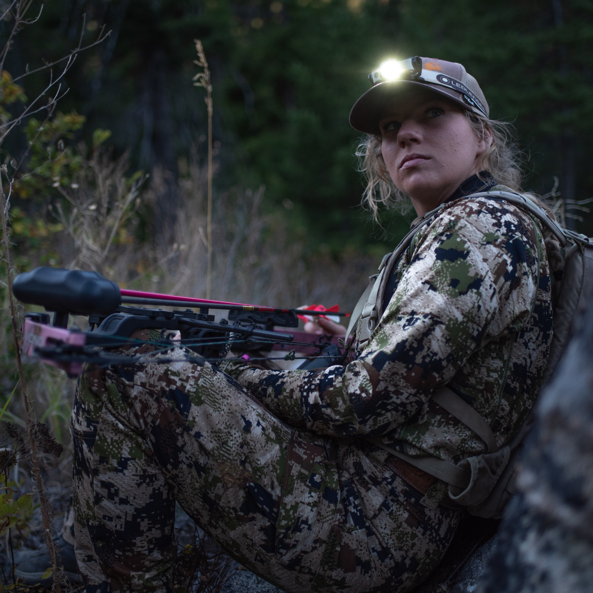 Person in camouflage outdoor gear wearing a Ledlenser MH4 Outdoor Headlamp on cap headlamp illuminated sitting with a bow in a forested area