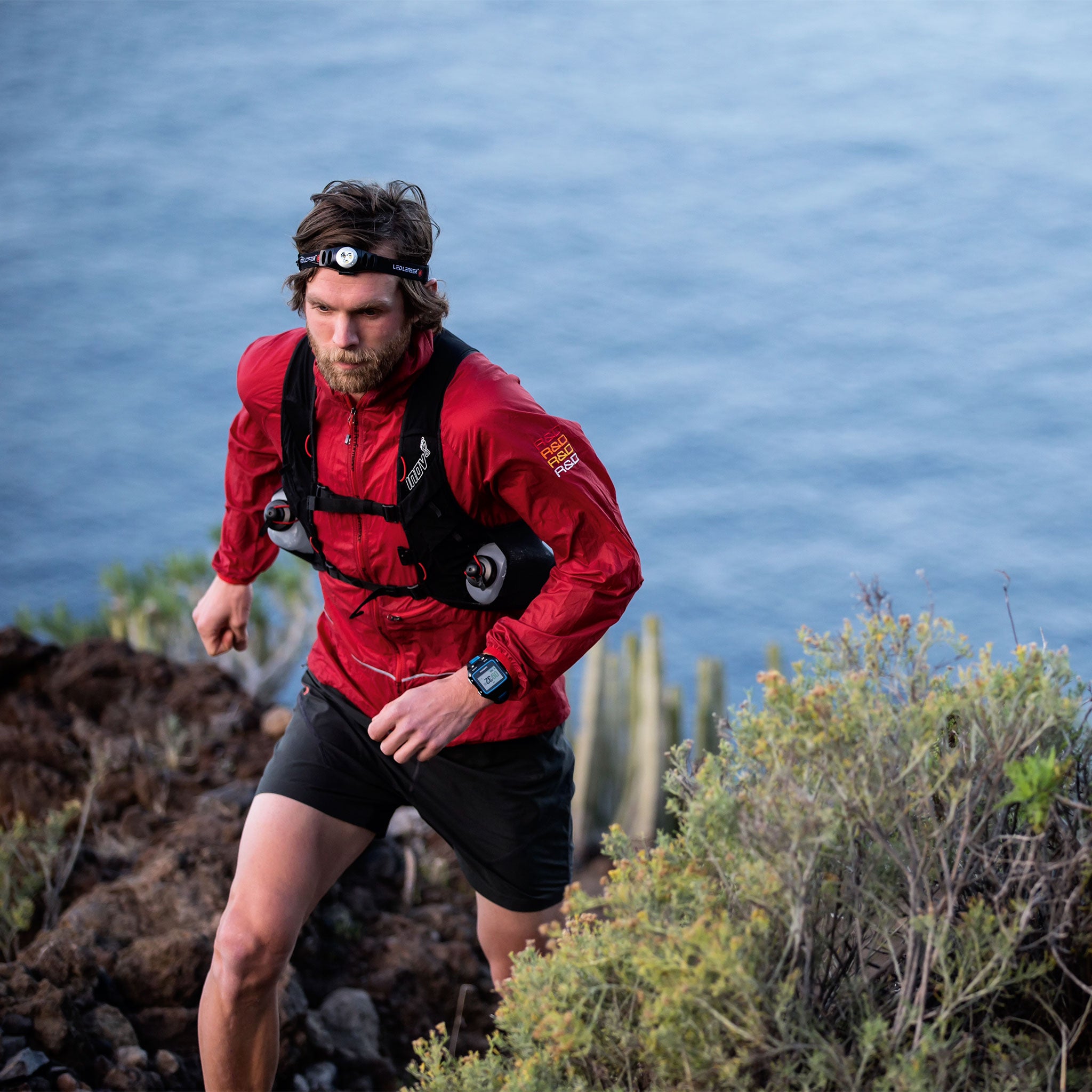 Ledlenser H3 2 Battery Operated Headlamp worn on a man's head during a coastal trail run in a red outdoor jacket with blue sea background