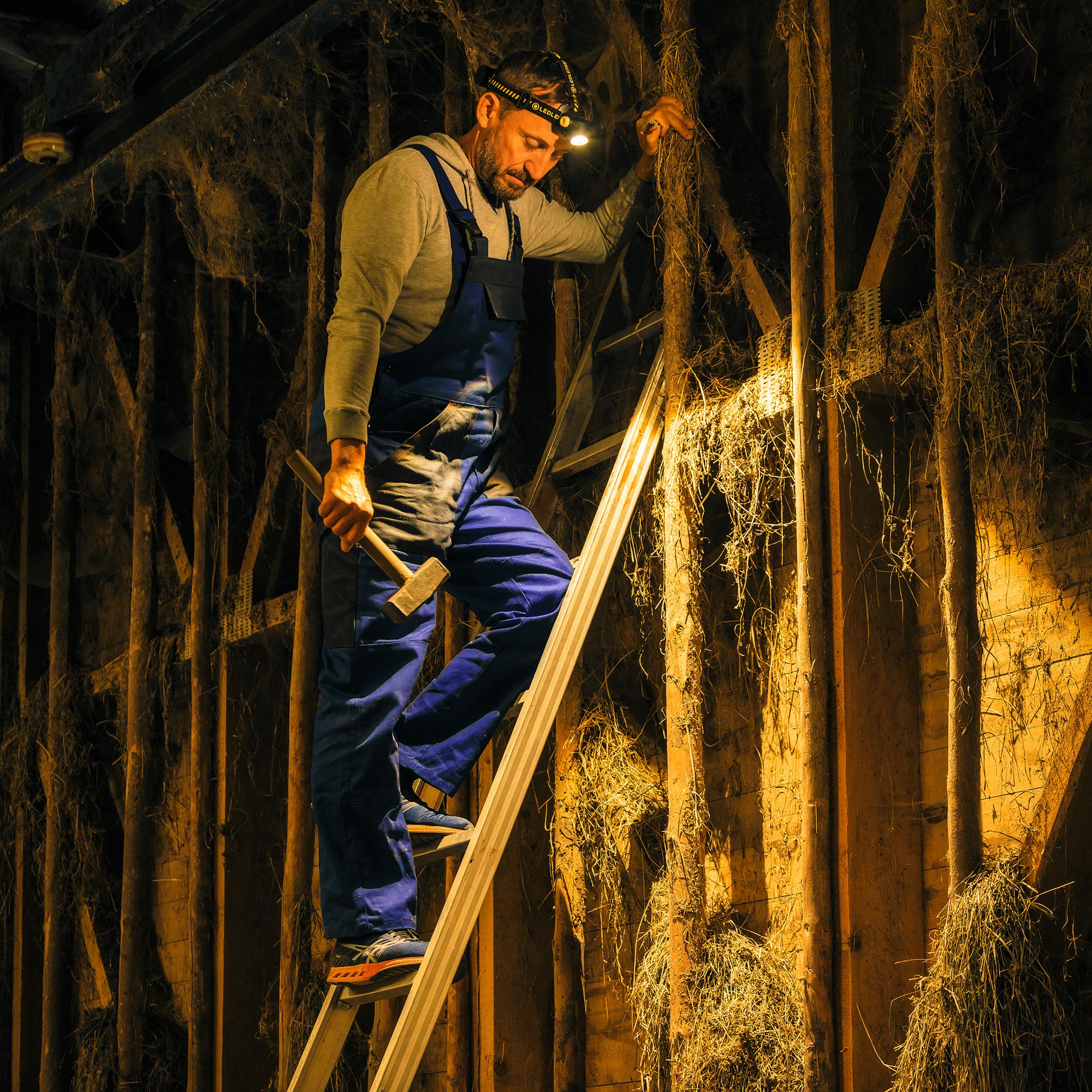 Ledlenser H7R Work Headlamp worn on the head by a man on a ladder in a dim workshop with a hammer showcasing professional work lighting