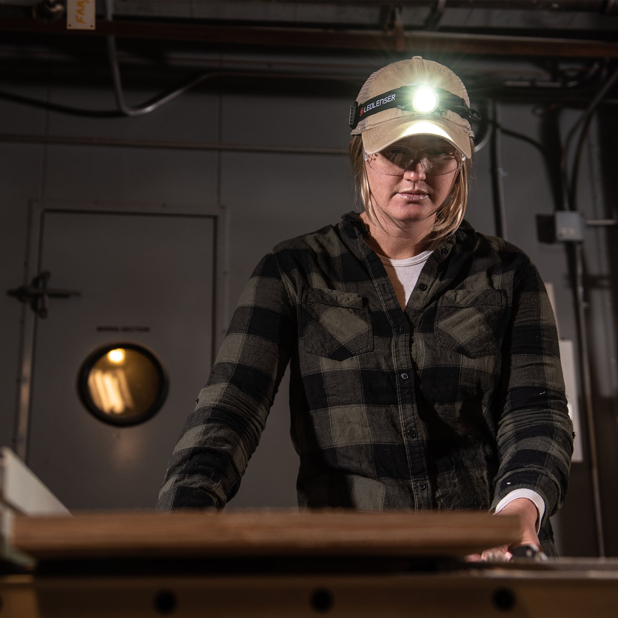 Ledlenser H8R Headlamp worn on a cap by a person in a workshop with the headlamp illuminated