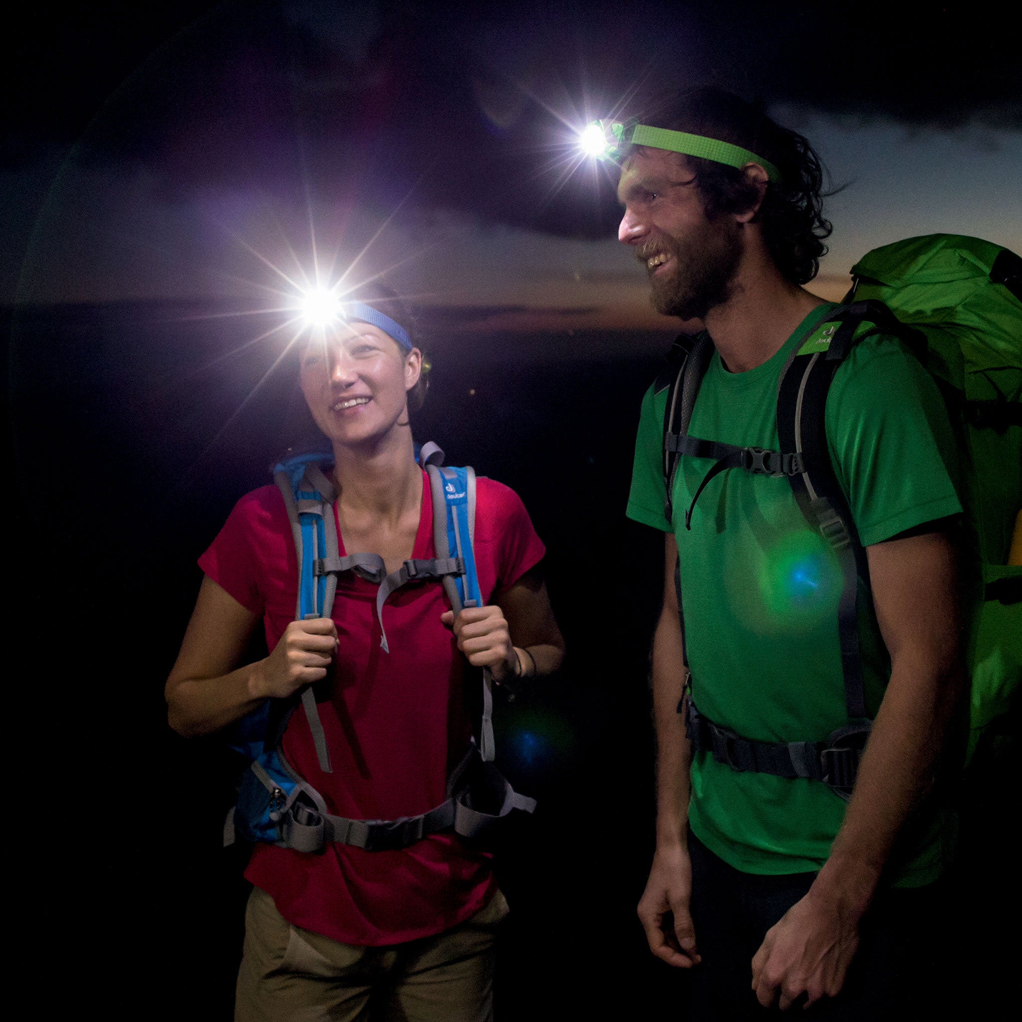 SEO3 Headlamp worn by a female hiker in red shirt at night; bright beam visible as a green backpacked male hiker stands nearby