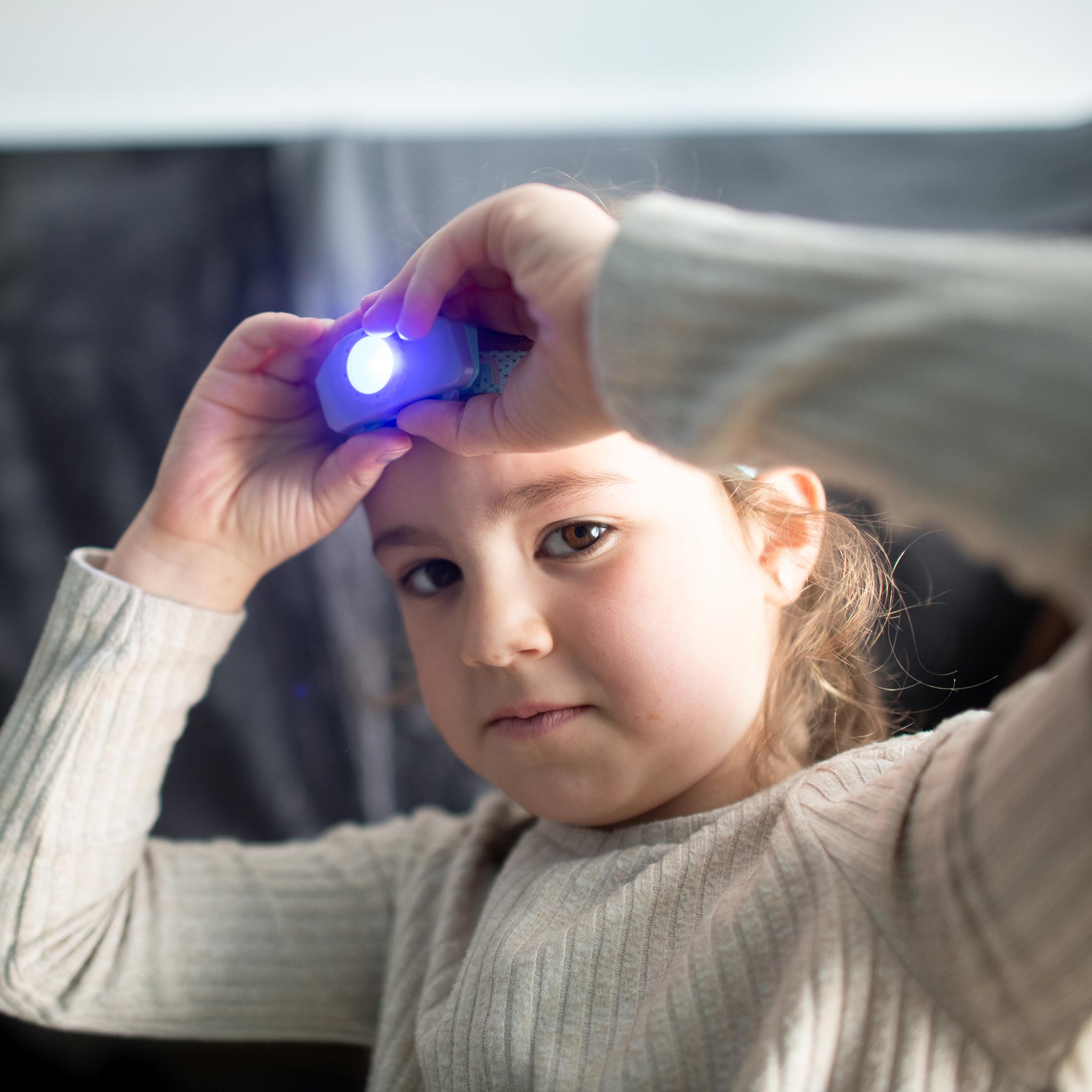 Kidled2 Headlamp held by a young girl with a blue illuminated LED at the front beige sweater visible