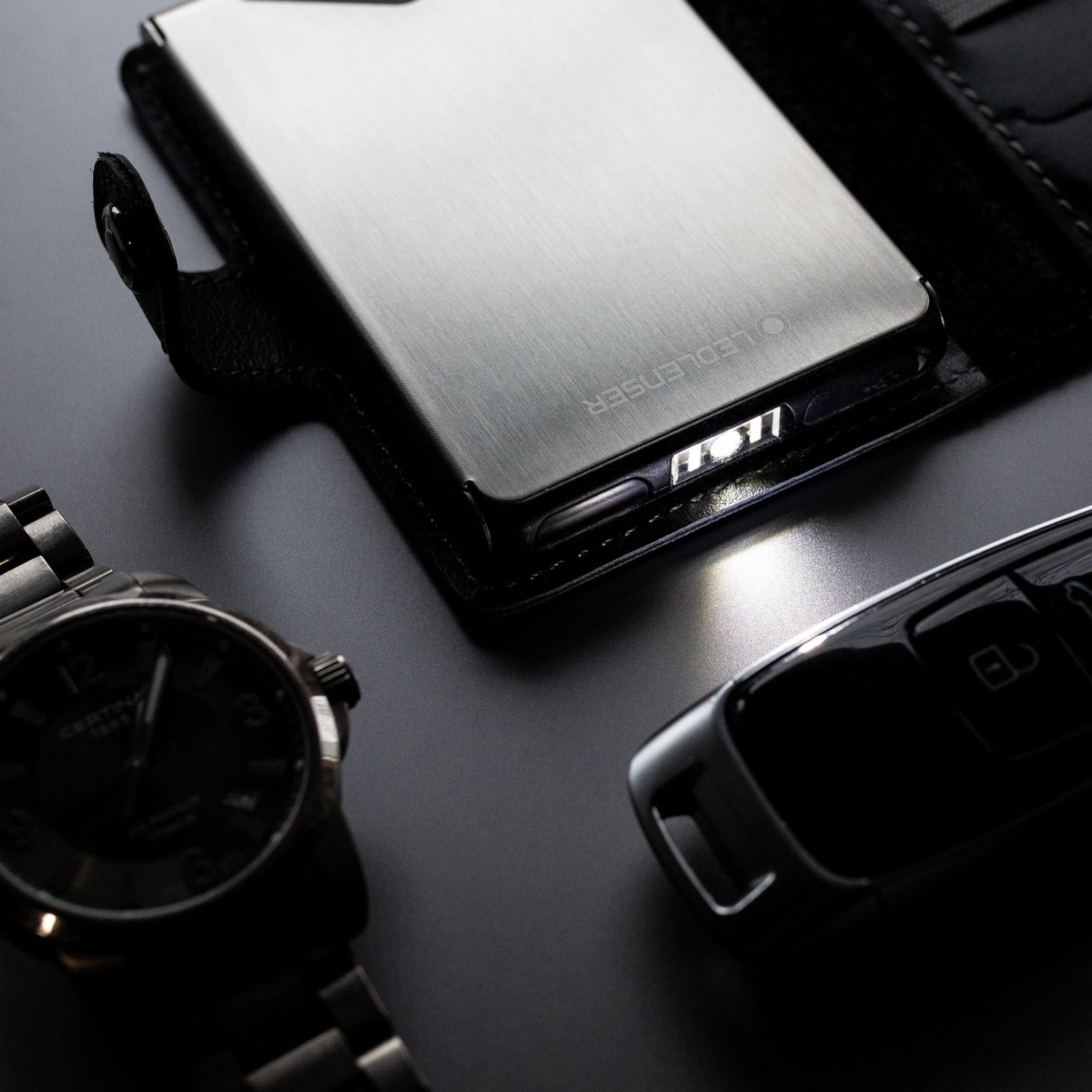 Ledlenser Lite Wallet® metal compact wallet with brushed aluminum finish closed with a small edge light visible shown beside a wristwatch and car key fob