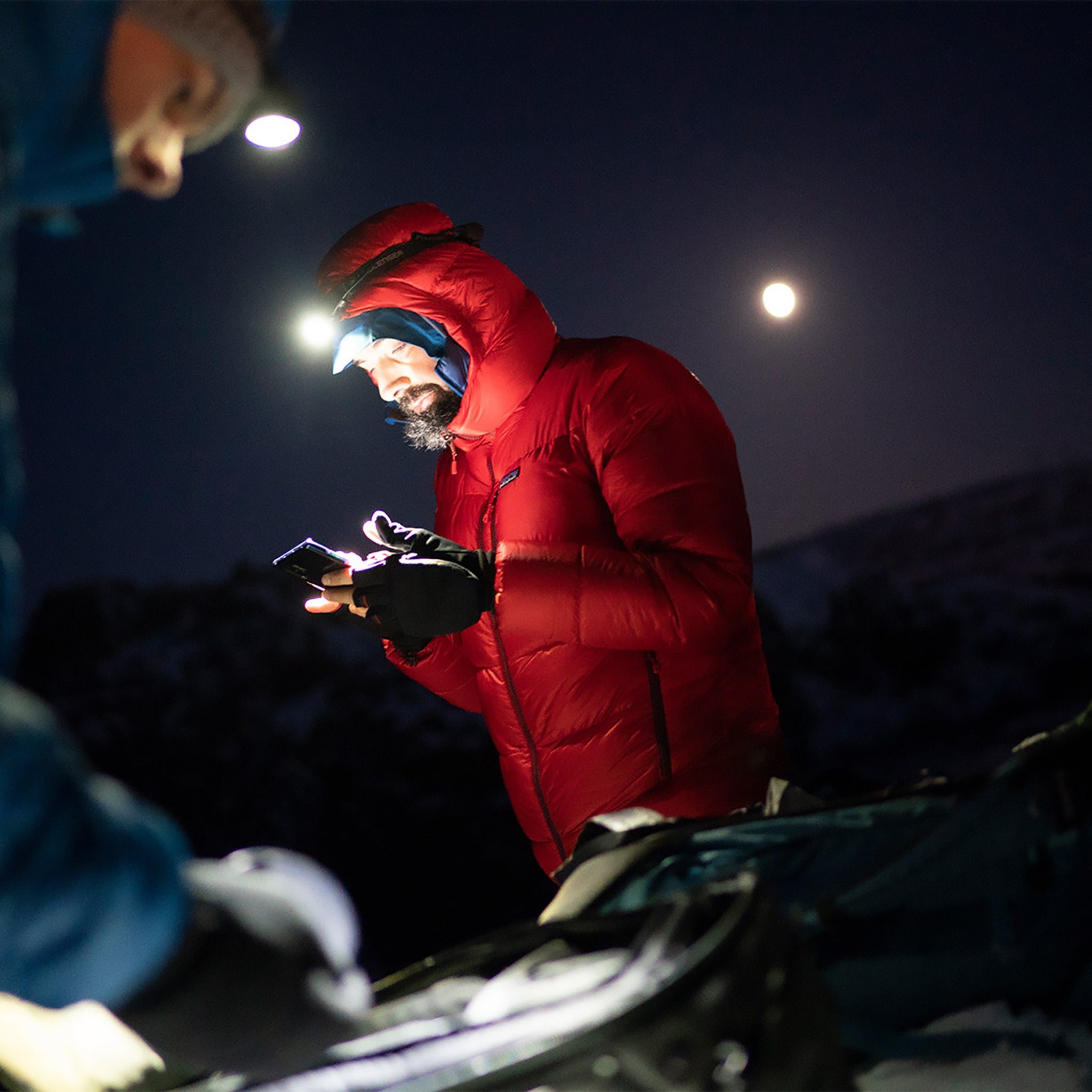 Ledlenser MH10 Headlamp worn by a man in a red outdoor jacket with a lit headlamp using a smartphone at night in a snowy outdoor setting
