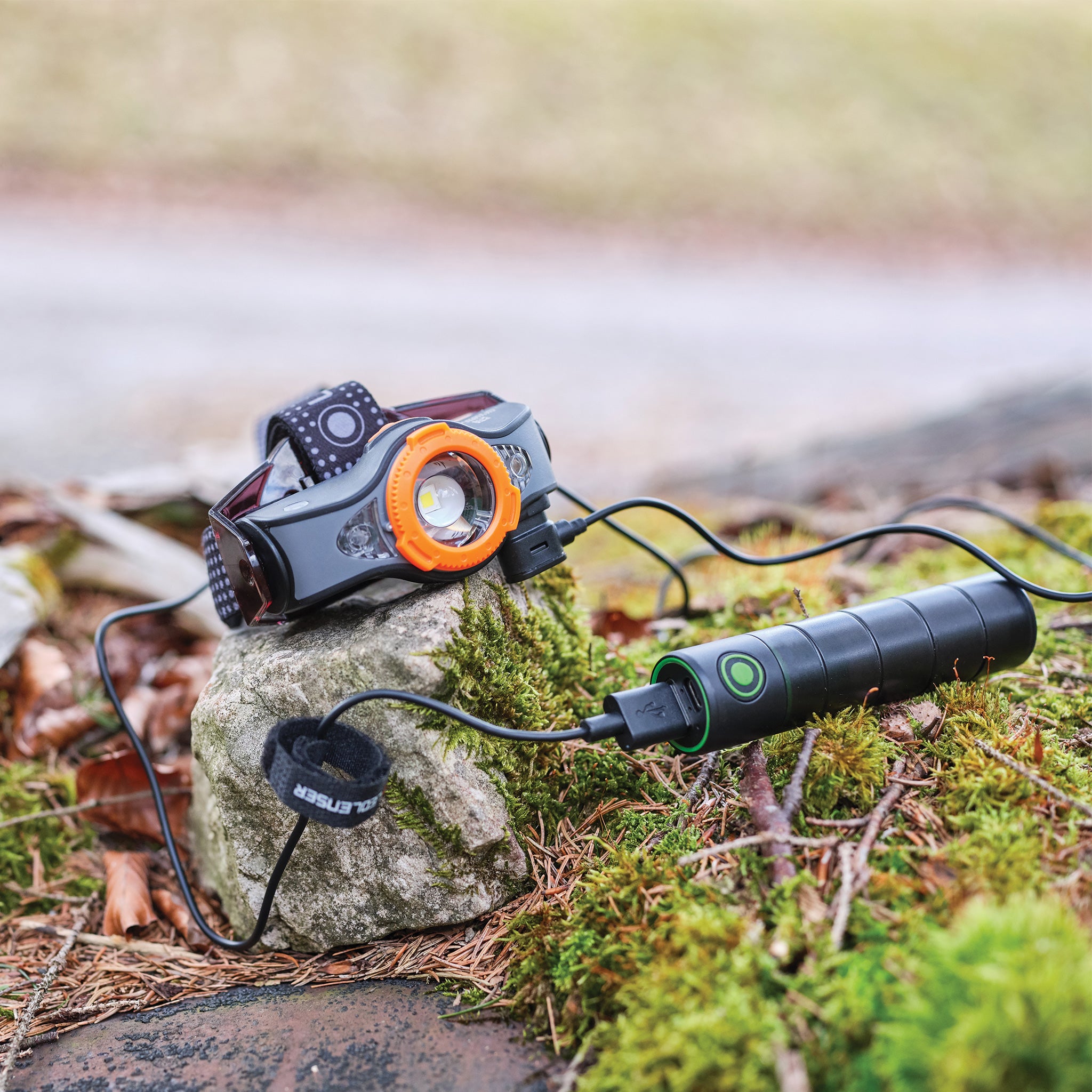 Flex3 Powerbank with headlamp attachment orange front ring and black cylindrical body on a mossy rock with a charging cable