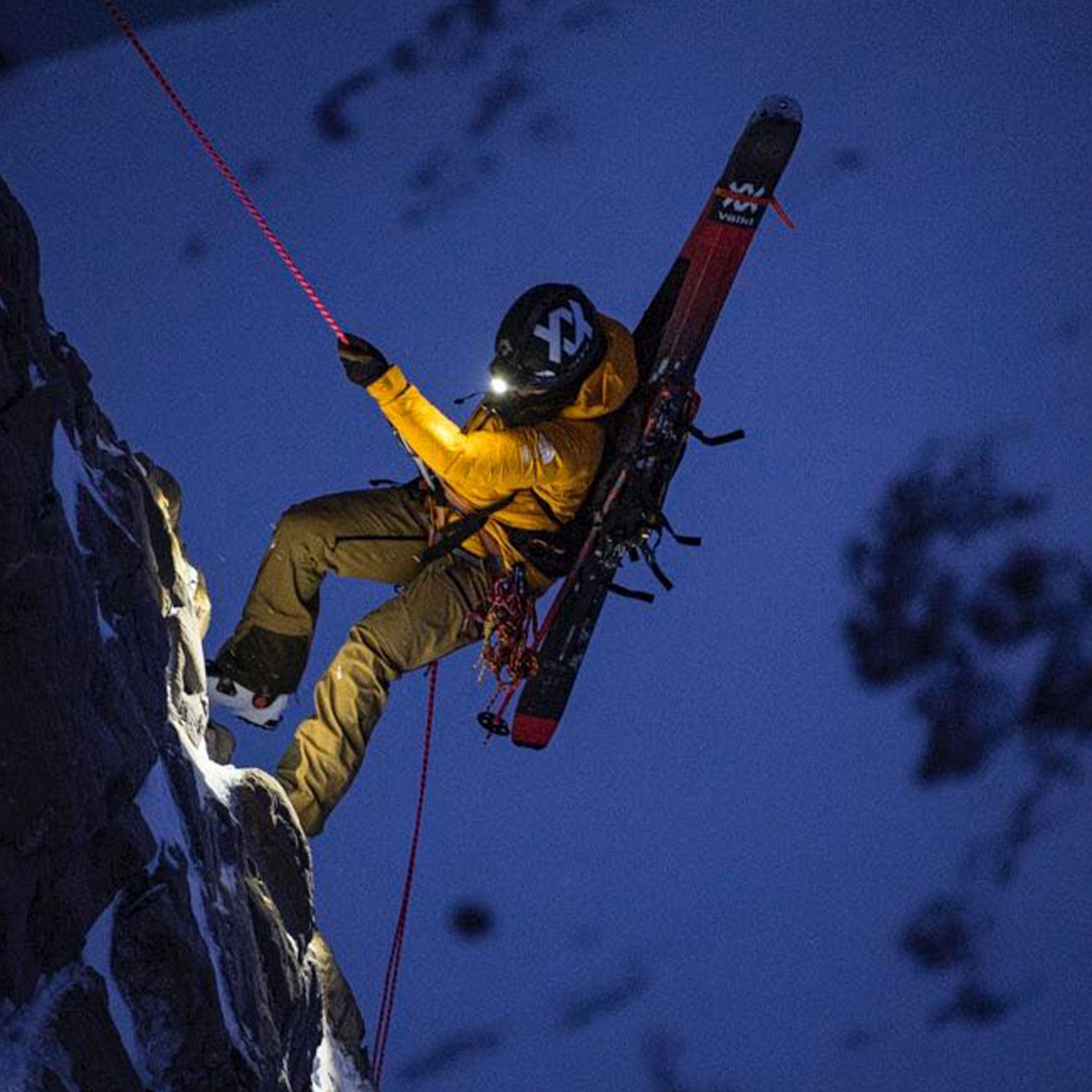 MH11 Outdoor Headlamp being used by a climber on a vertical rock face at night; wearer in a yellow jacket and helmet with rope and gear headlamp illuminated