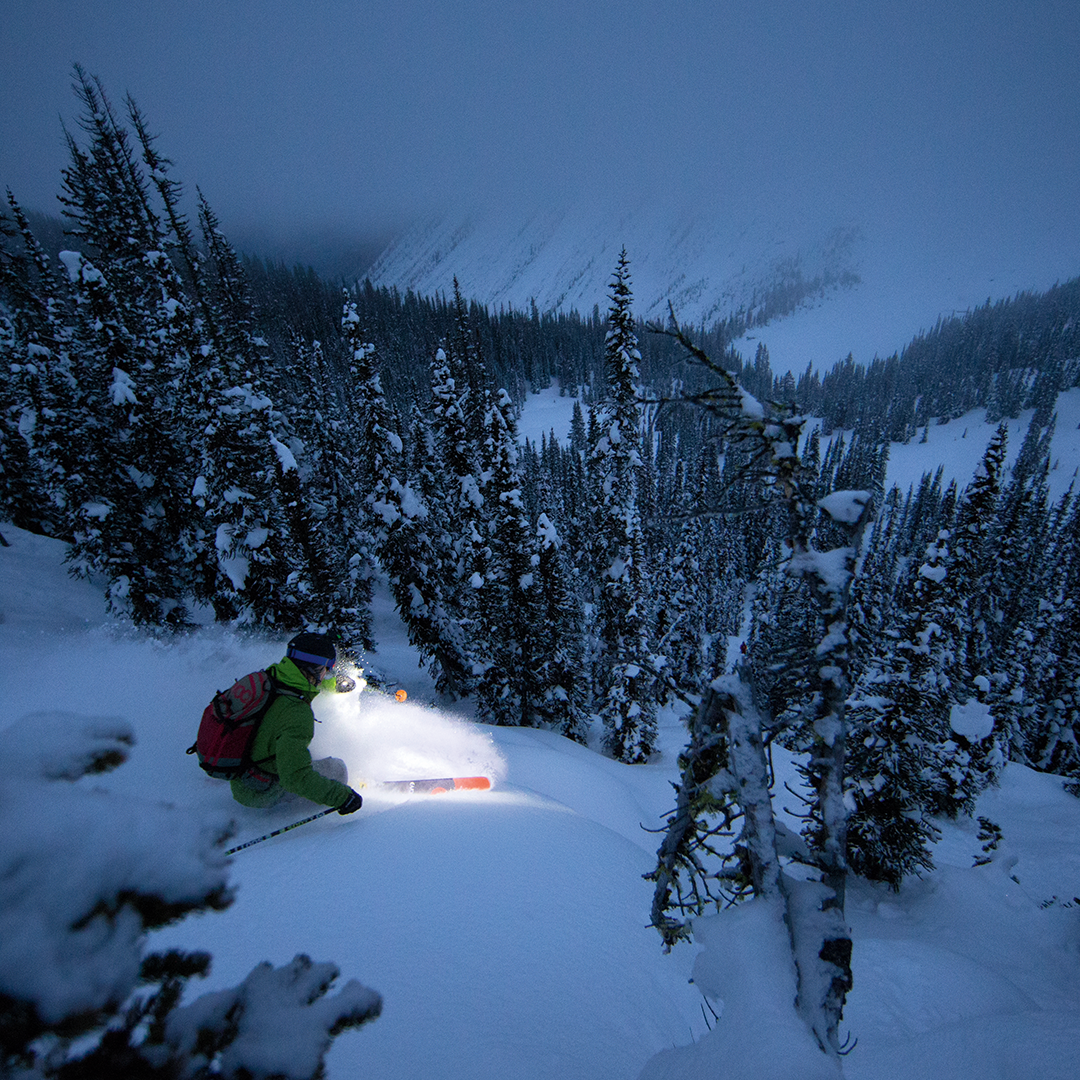 Ledlenser XEO19R Rechargeable Headlamp worn by a backcountry skier on a snowy slope with pine trees at dusk