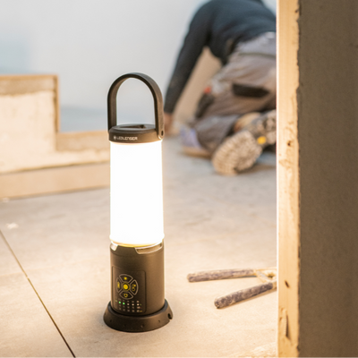 AL10R Work Lantern by Ledlenser upright on a concrete floor with illuminated cylindrical light black base with control panel and curved carrying handle; construction site backdrop