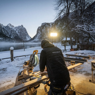 AF8R Work Floodlight in an outdoor snowy workshop; a worker in a jacket uses a miter saw at a wooden workbench with a mounted LED floodlight