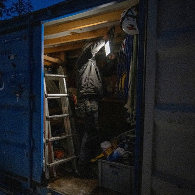 AF8R Work Floodlight in use inside a dark storage trailer with a person in a jacket reaching overhead a ladder and tools nearby