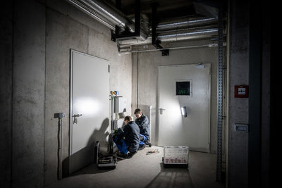 AF8R Work Floodlight in an industrial room with two technicians kneeling beside electrical panels under overhead pipes