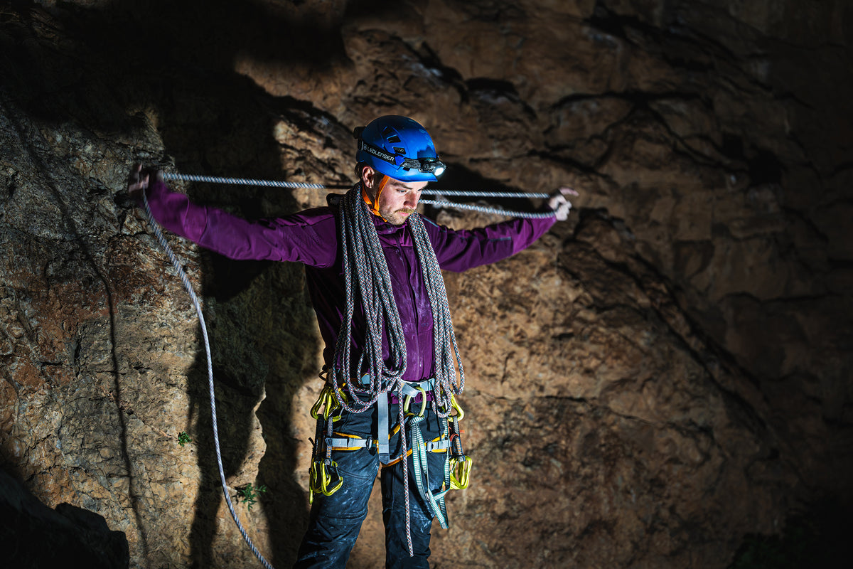 A climber wearing a blue helmet and purple jacket adjusts a rope in a dark cave, illuminated by a Ledlenser H19R Pro headlamp.