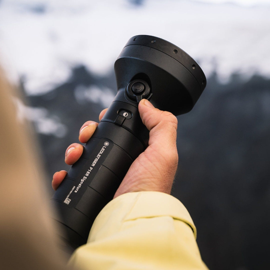 Ledlenser P18R Signature Torch held in hand showing its large round head and black cylindrical body against an outdoor blurred background