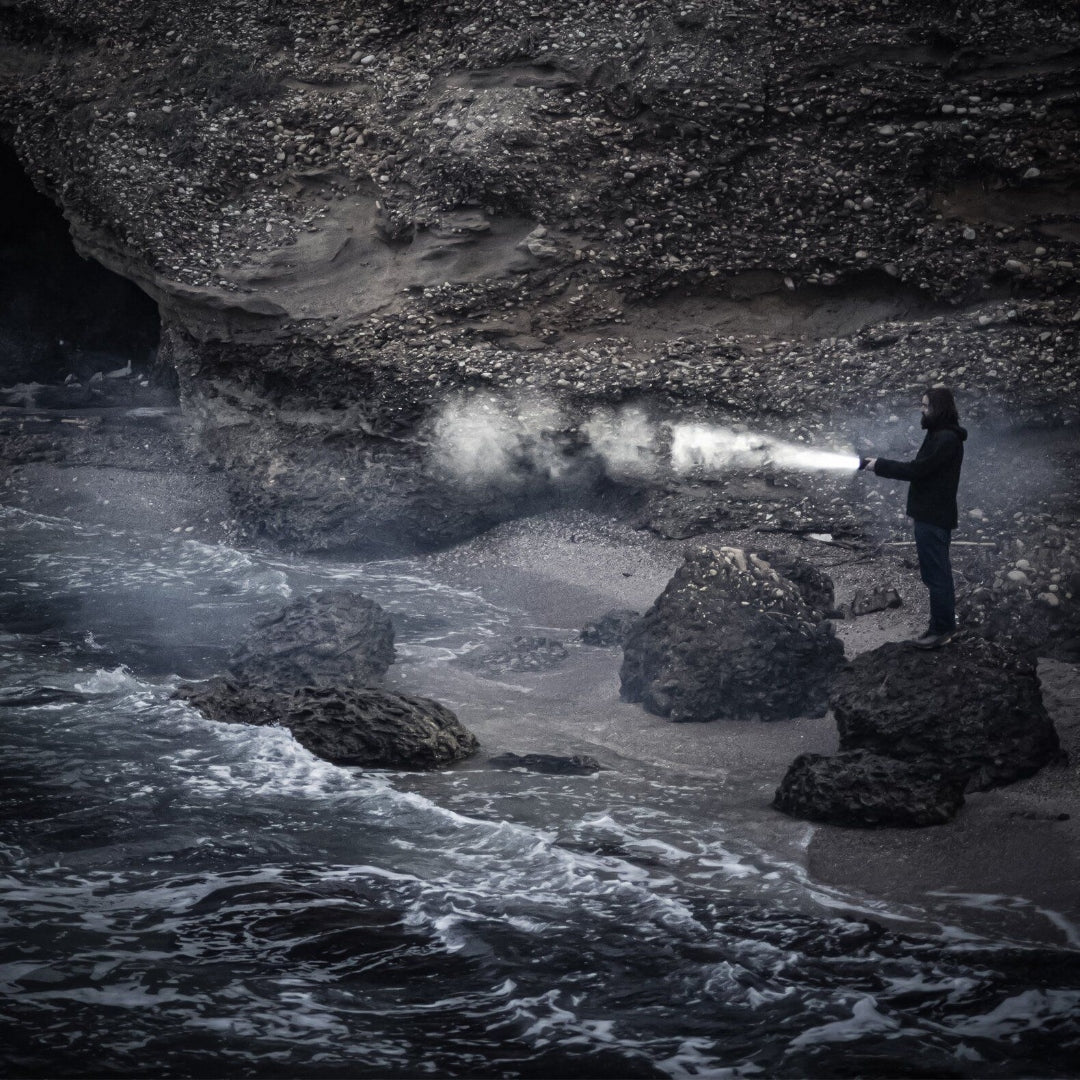 P18R Signature Torch held by a person on a dark rocky shoreline beam projecting across mist toward the water