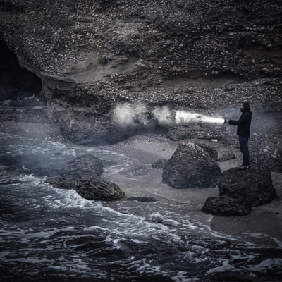 P18R Signature Torch held by a person on a dark rocky shoreline beam projecting across mist toward the water