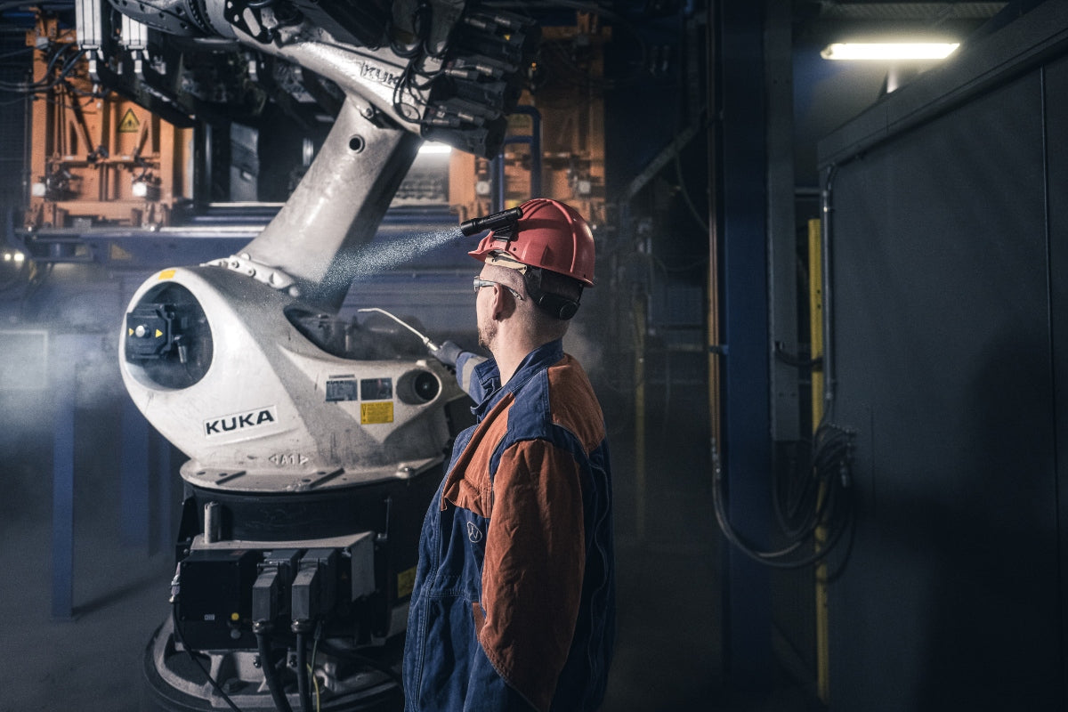 P5R Torch industrial scene: worker in orange blue coveralls and red helmet near a KUKA robotic arm in a dim workshop