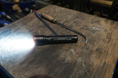 Ledlenser P7R Torch on a workbench with a bright beam to the left black cylindrical flashlight with a charging cable and green indicator LEDs visible