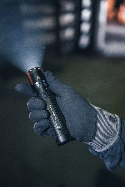Ledlenser P7R Torch held in a gloved hand with beam projecting forward black cylindrical body with a red ring at the head and visible branding
