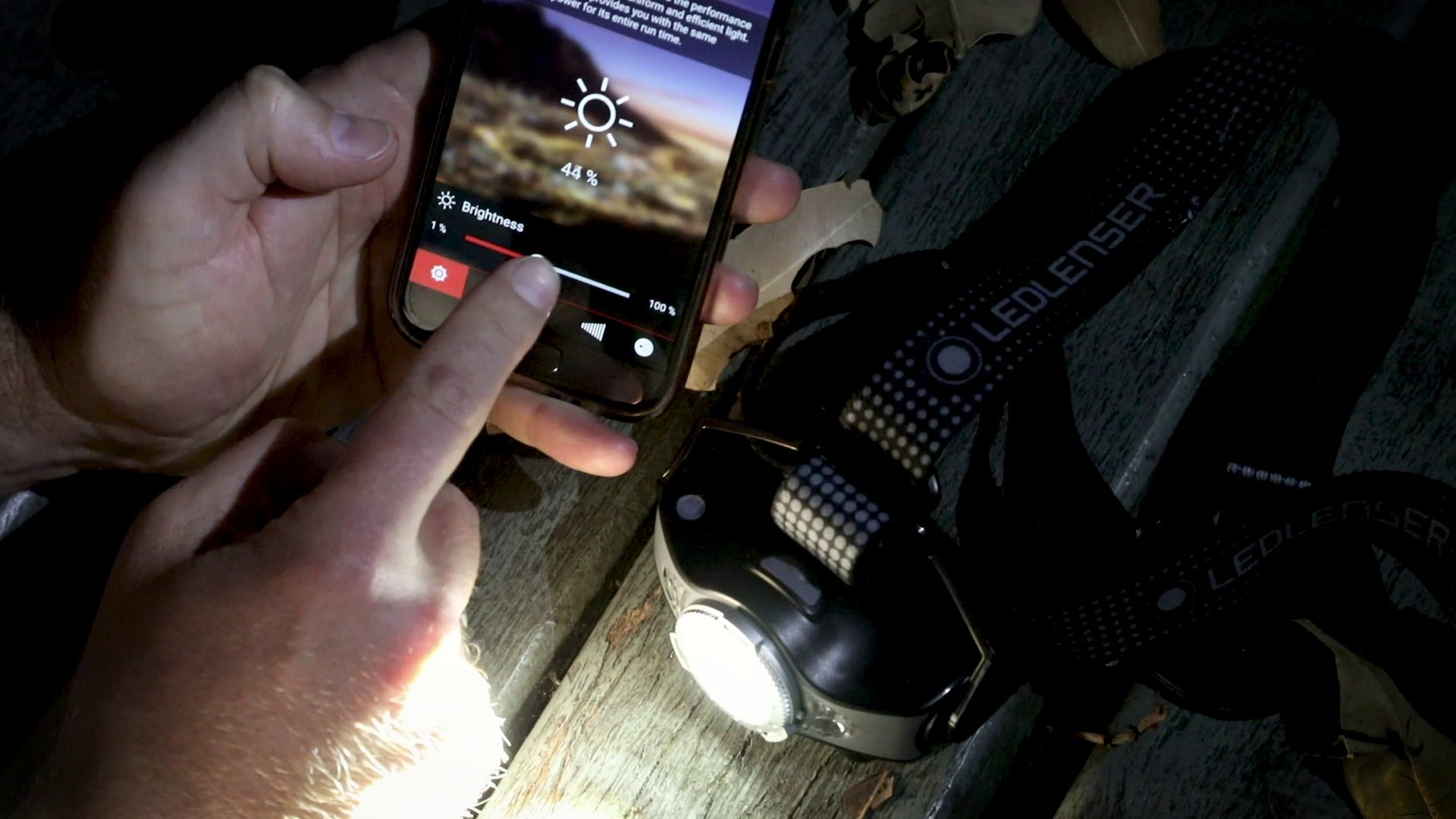 MH11 Blue Outdoor Headlamp with blue housing and strap shown on a wooden surface next to a smartphone displaying brightness controls
