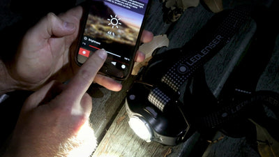 MH11 Blue Outdoor Headlamp with blue housing and strap shown on a wooden surface next to a smartphone displaying brightness controls