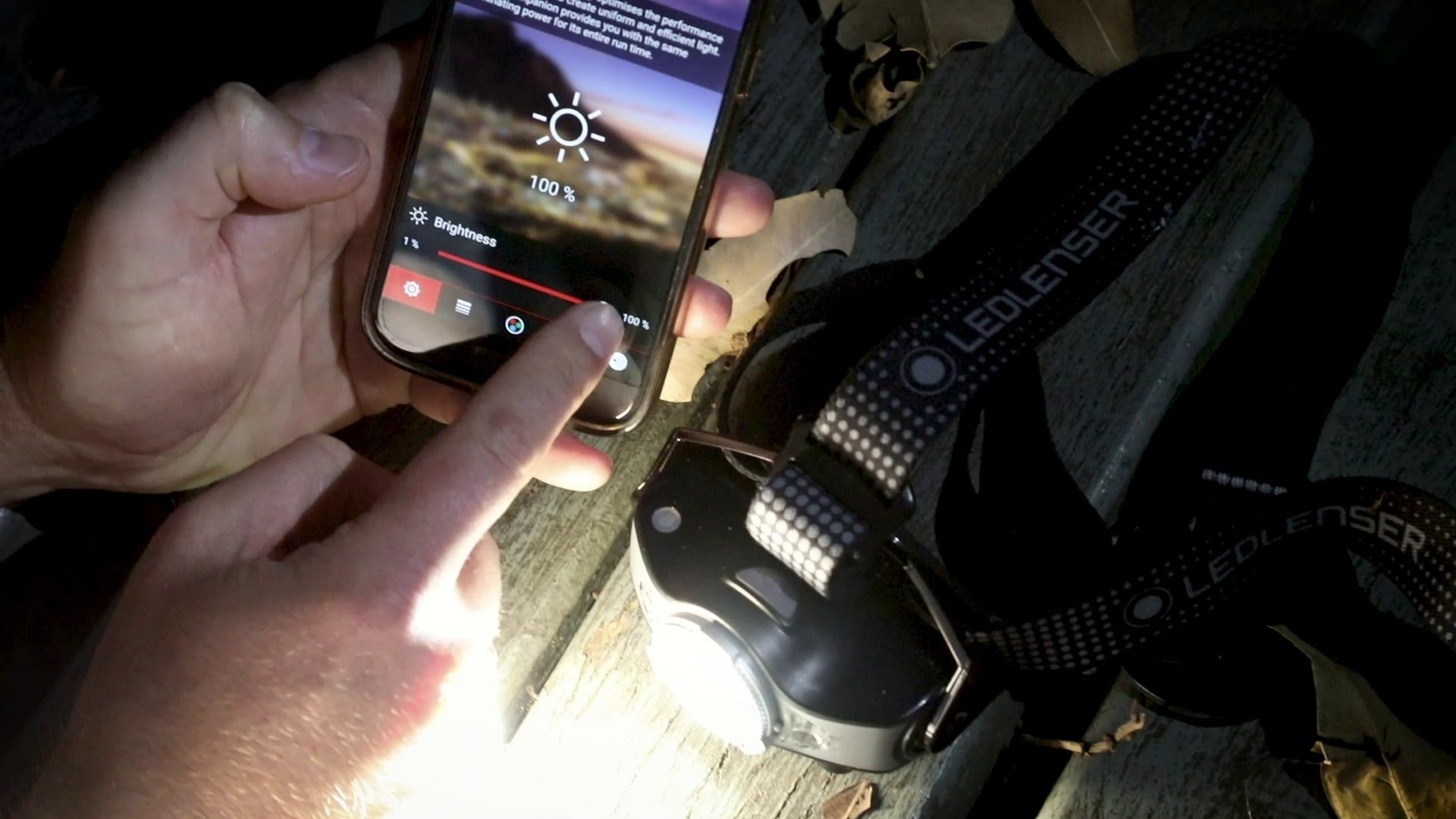 MH11 Blue Outdoor Headlamp by Ledlenser on a wooden surface next to a smartphone showing brightness controls