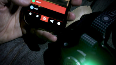 MH11 Blue Outdoor Headlamp held in hand with a green light beam; smartphone screen shows color options green and blue