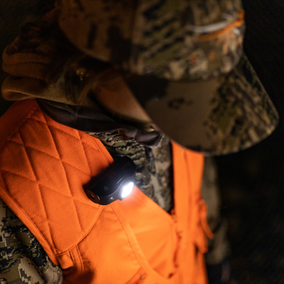 MH8 Powercase Combo headlamp clipped to an orange safety vest worn by a person in camouflage gear headlamp emitting bright white light