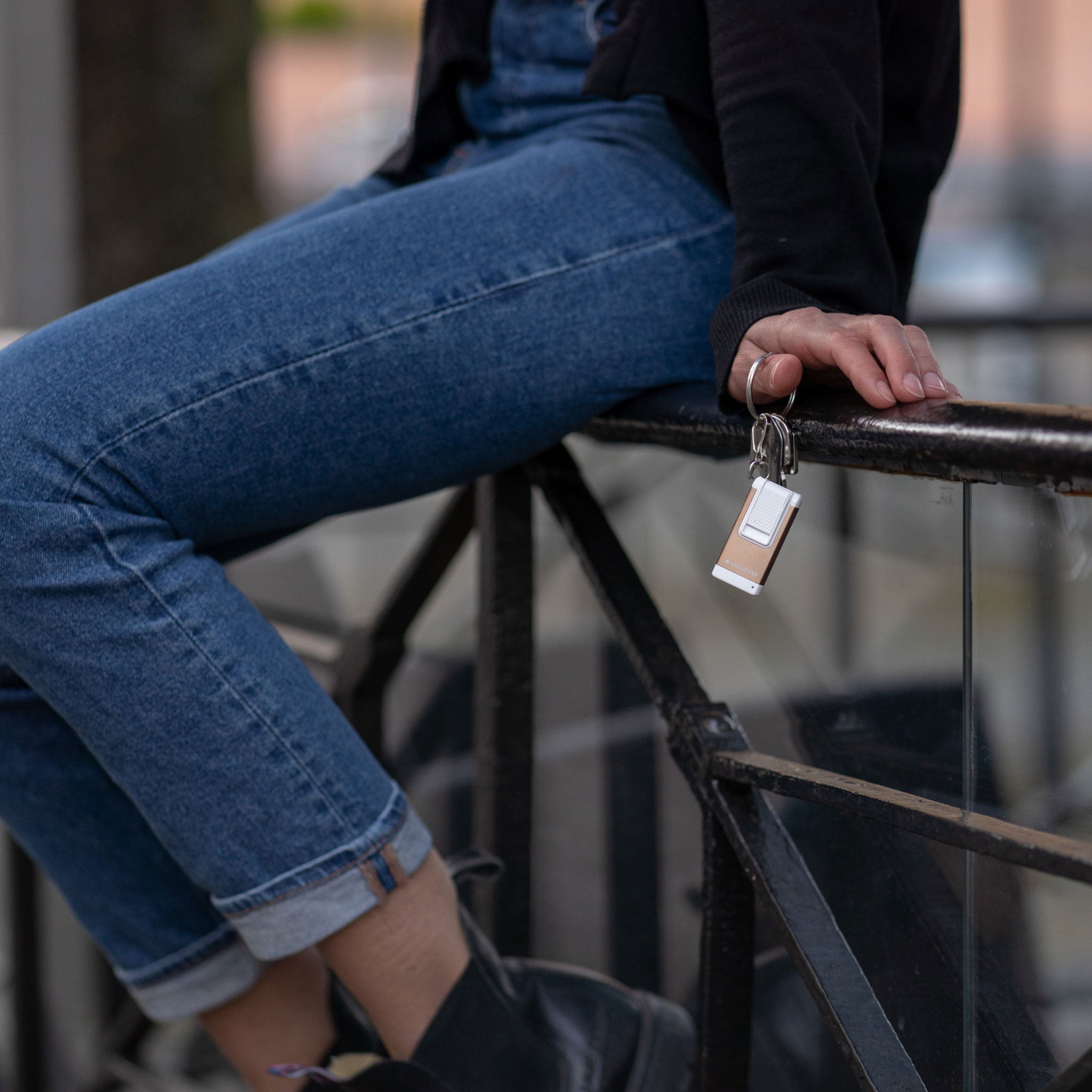 Ledlenser K4R keychain light attached to a keyring beige body with white top held by a person sitting on a railing in blue jeans