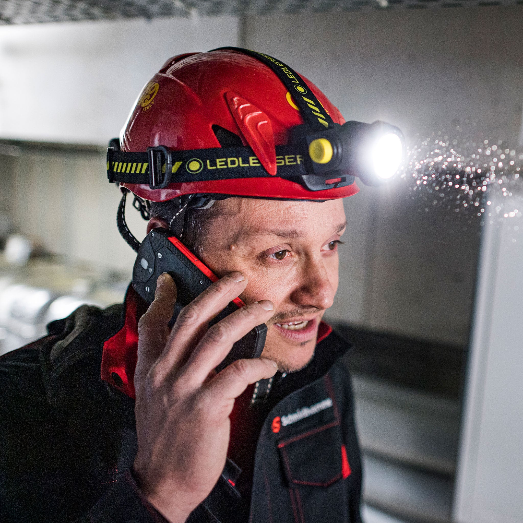 Ledlenser H15R Work Headlamp mounted on a red helmet with strap; headlamp illuminated worker on phone in an industrial setting