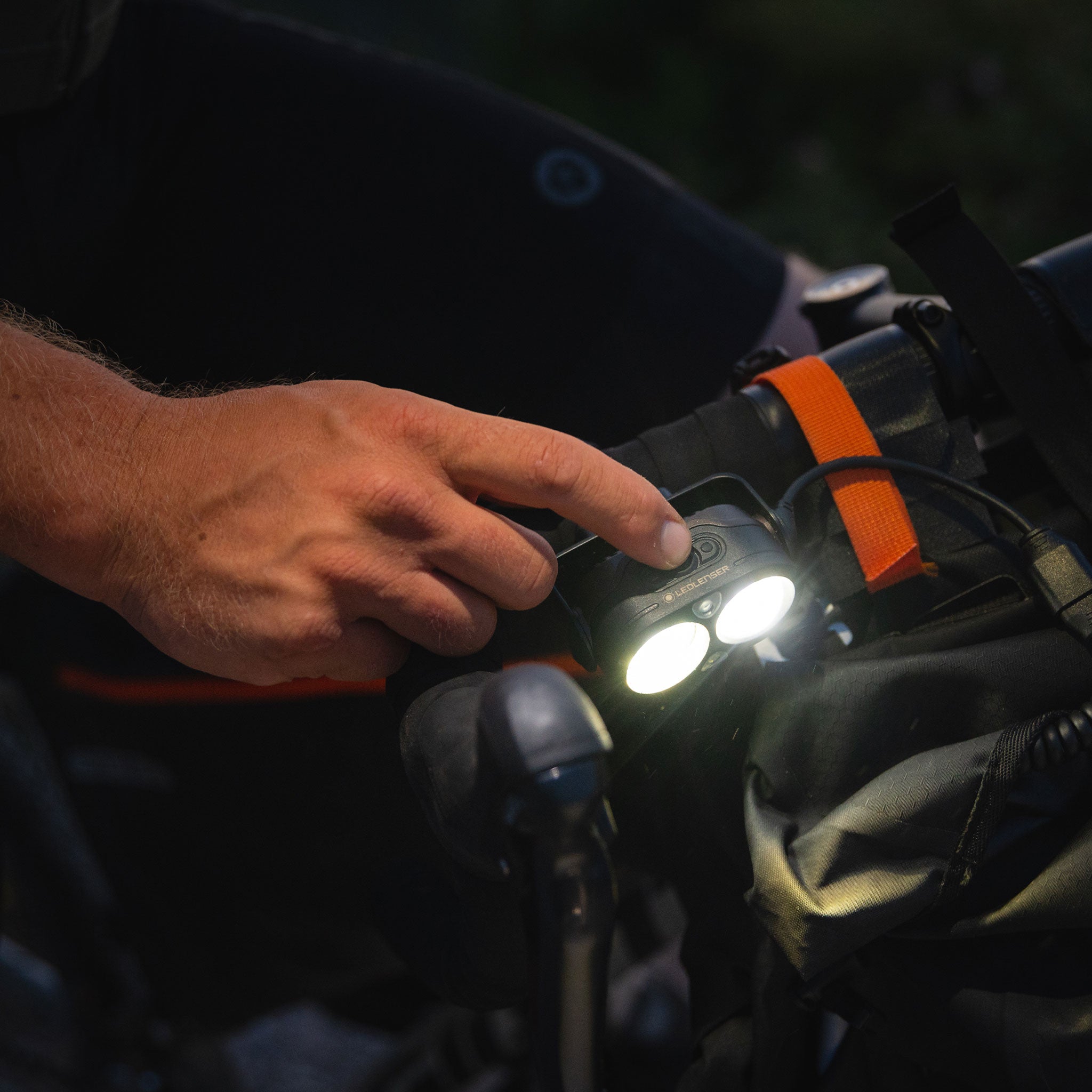 Ledlenser H19R Signature Headlamp mounted on a backpack with an orange strap hand pressing the power button and dual LEDs illuminated