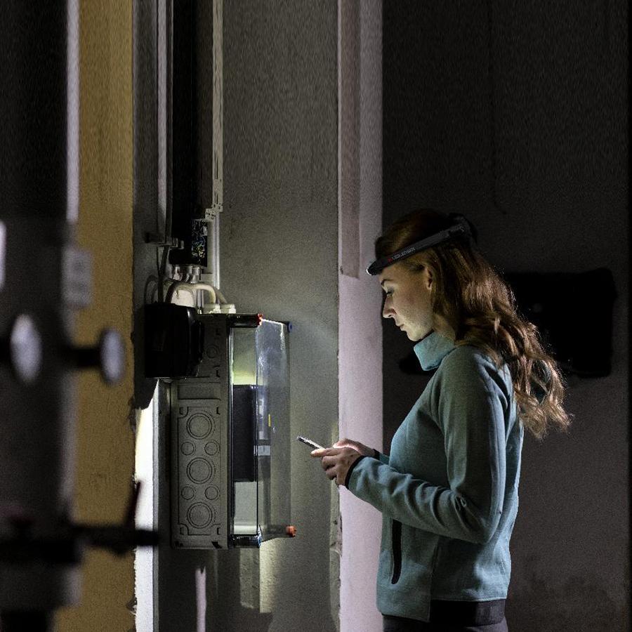 H4R Rechargeable Headlamp worn by a woman in a dim lab headlamp on examining a smartphone beside a wall mounted control panel