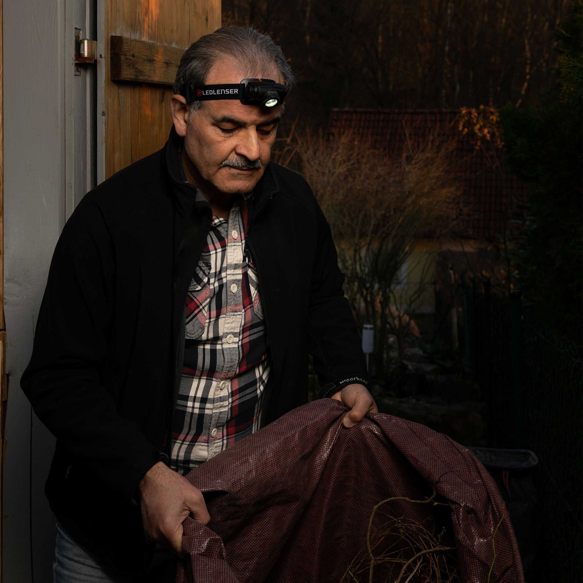 Ledlenser H5 Core Headlamp worn by a man outdoors near a shed headlamp visible on his forehead as he holds a bag