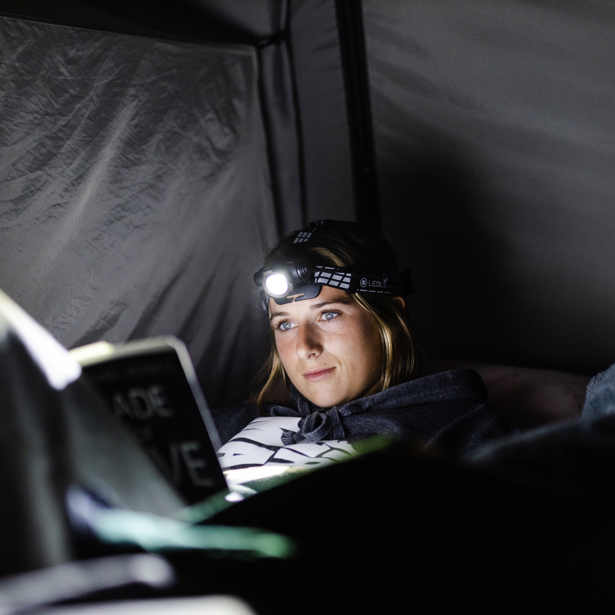 Ledlenser H7R Signature Headlamp worn on head by a woman inside a tent with the light on while reading a book