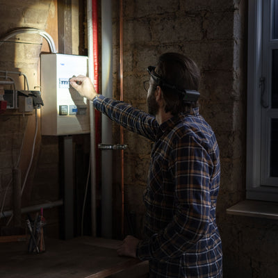 H7R Core Headlamp worn by a man in a plaid shirt guiding a finger toward an electrical panel in a dim workshop basement