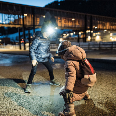 Kidled2 Headlamp worn by child in brown jacket with backpack outdoors at night while another person nearby wears a bright headlamp