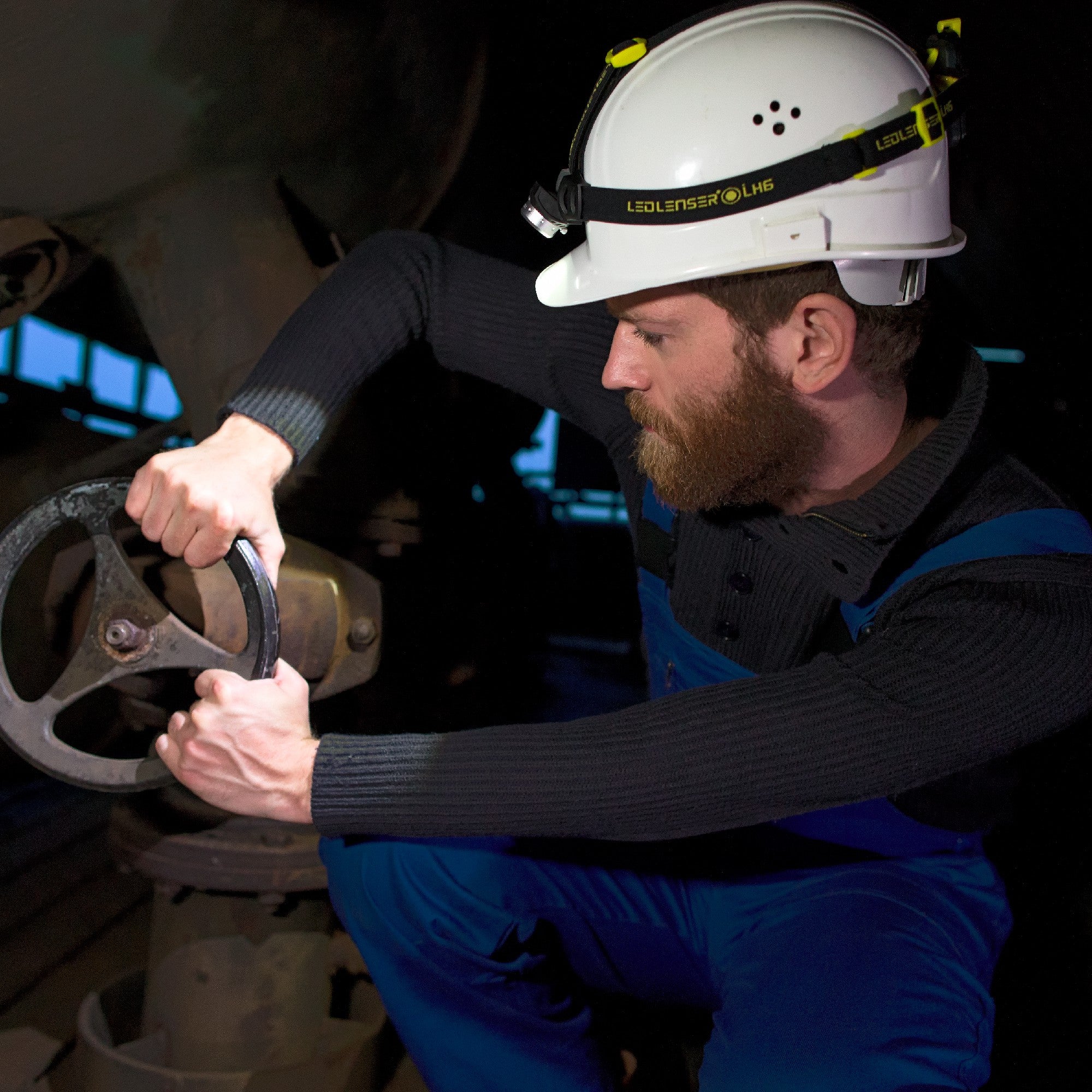 iH6R Rechargeable Headlamp worn on a white safety helmet by a bearded worker in a dark industrial setting