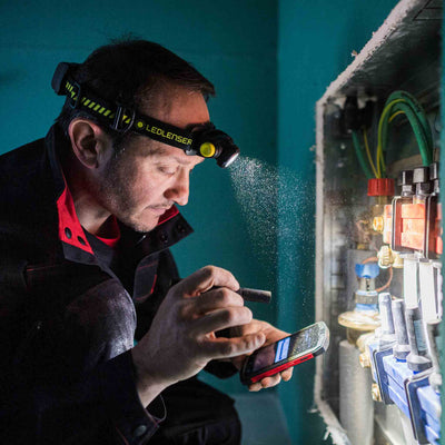 P2 Battery Operated Torch headlamp on a man’s head with yellow accents inspecting an electrical panel with sparks and holding a smartphone