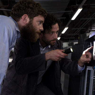 i7 Torch being used by two professionals inspecting a rack mounted control panel in a dim workshop