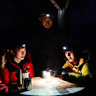 MH5 Outdoor Headlamp worn by a person at night with a group gathered around a lit table and headlamps illuminating faces