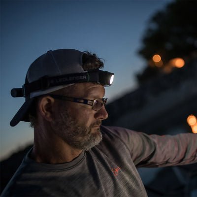 MH10 Headlamp worn on a man’s head outdoors at dusk; compact black headlamp emitting light with Ledlenser on the strap