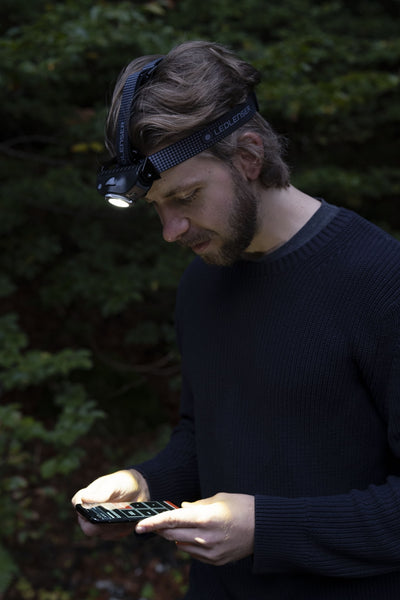 MH11 Blue Outdoor Headlamp worn on head by man outdoors strap with Ledlenser branding while looking at a smartphone