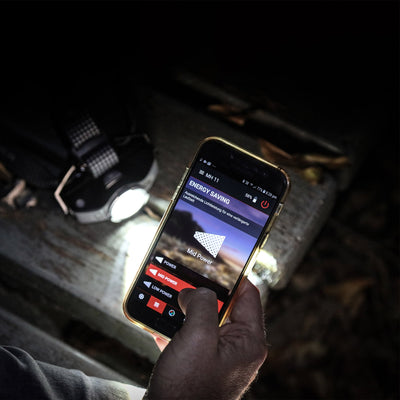 MH11 Blue Outdoor Headlamp shown with a headlamp strap on a wooden surface while a hand holds a smartphone displaying the MH11 energy saving screen