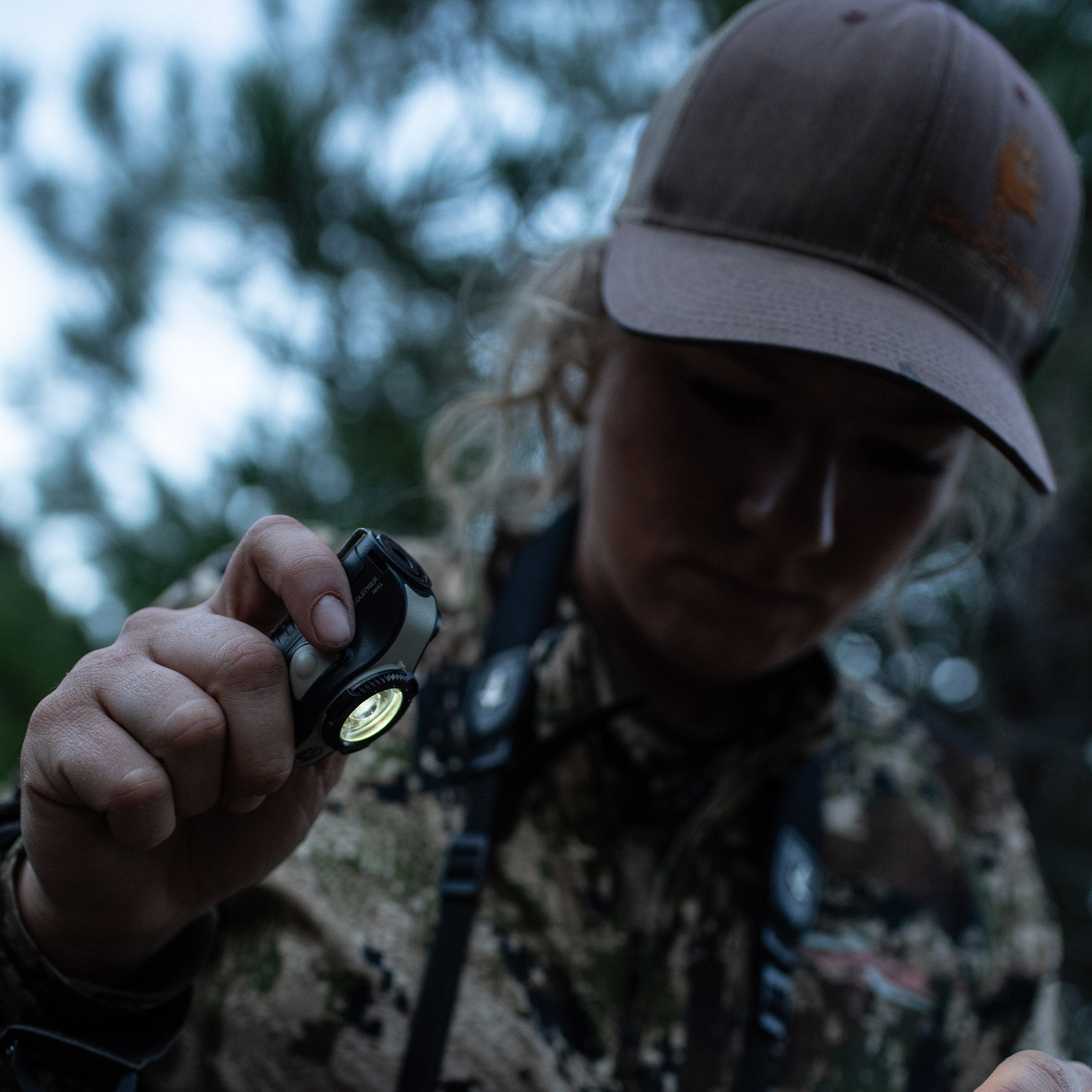 MH4 Outdoor Headlamp held by a person wearing camouflage clothing and a cap with the light on in an outdoor setting