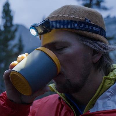 MH5 Outdoor Headlamp worn on a man's forehead with blue light Ledlenser strap outdoors while drinking from a mug
