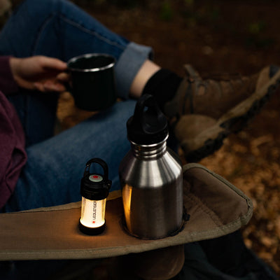 ML4 Warm Light Lantern shown on a camping sleeve next to a metal water bottle with a warm glow and a person holding a mug in the background
