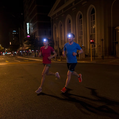 NEO6R Rechargeable Headlamp worn by a man and a woman running at night on an urban street; blue and red shirts headlamps lit