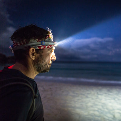 NEO6R Rechargeable Headlamp worn on a headband by a man at night on a beach with the beam projecting forward