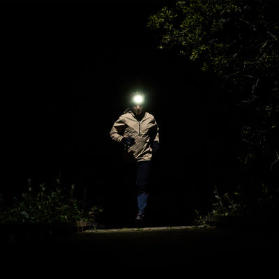 NEO9R Running Headlamp worn on the head of a runner wearing a beige jacket on a dark night trail with illumination from the headlamp